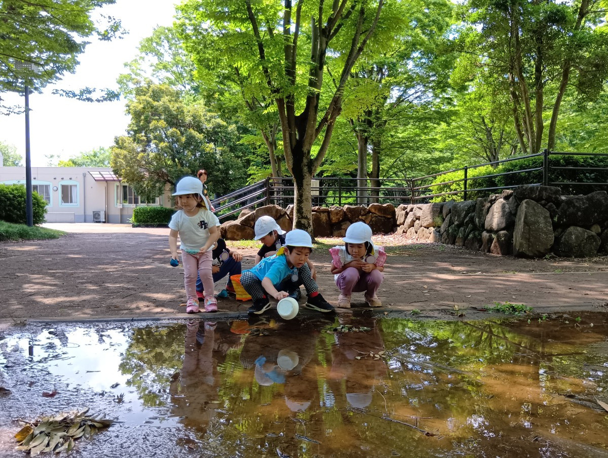 公園の水たまりの淵で、子どもたちはスコップやカップを手に持ち水面を見ている