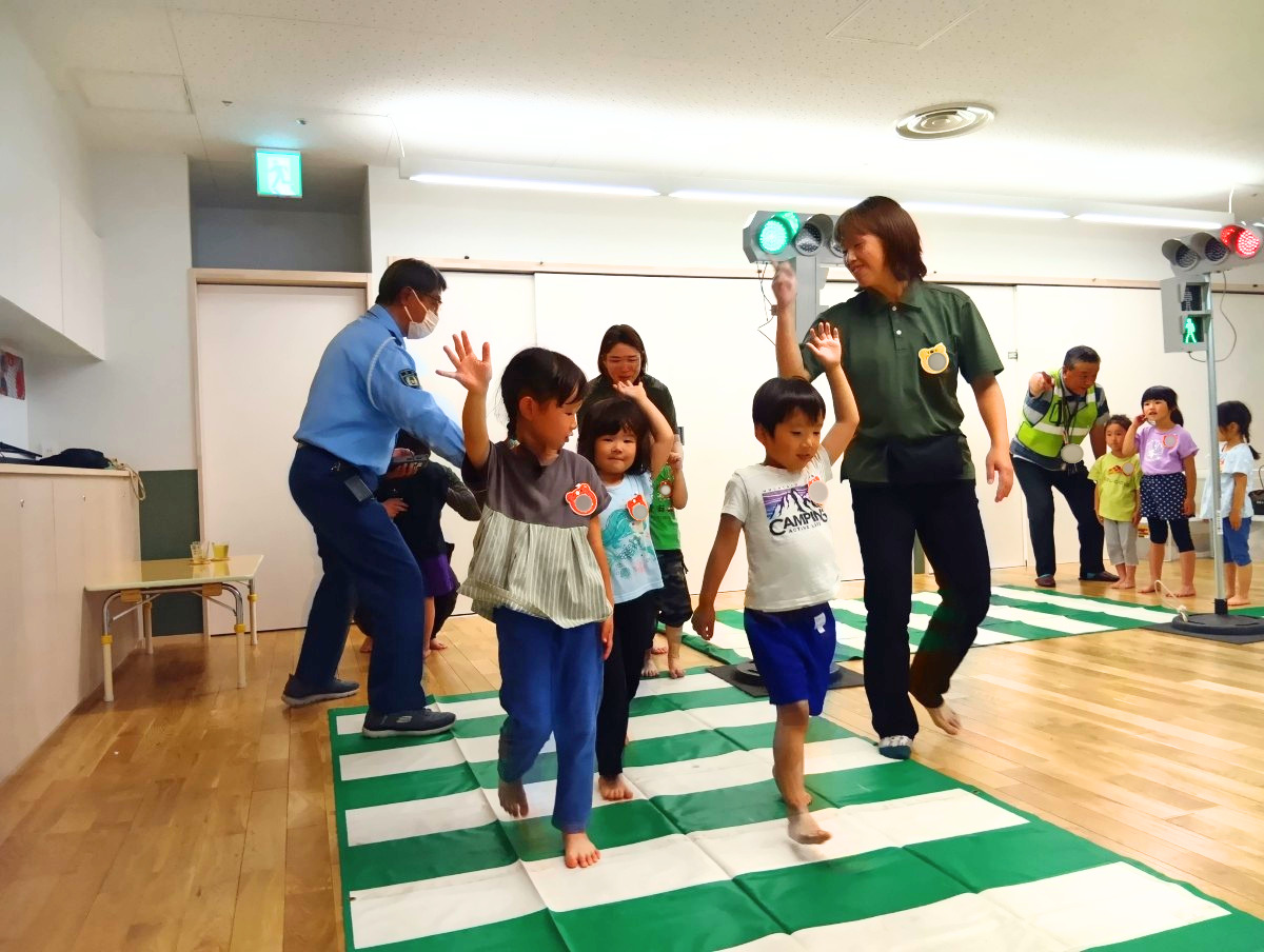 室内では、床に横断歩道のシートと信号機が設置されおり、信号が青点灯になり横断歩道の上を子どもたちが手をあげて歩いている