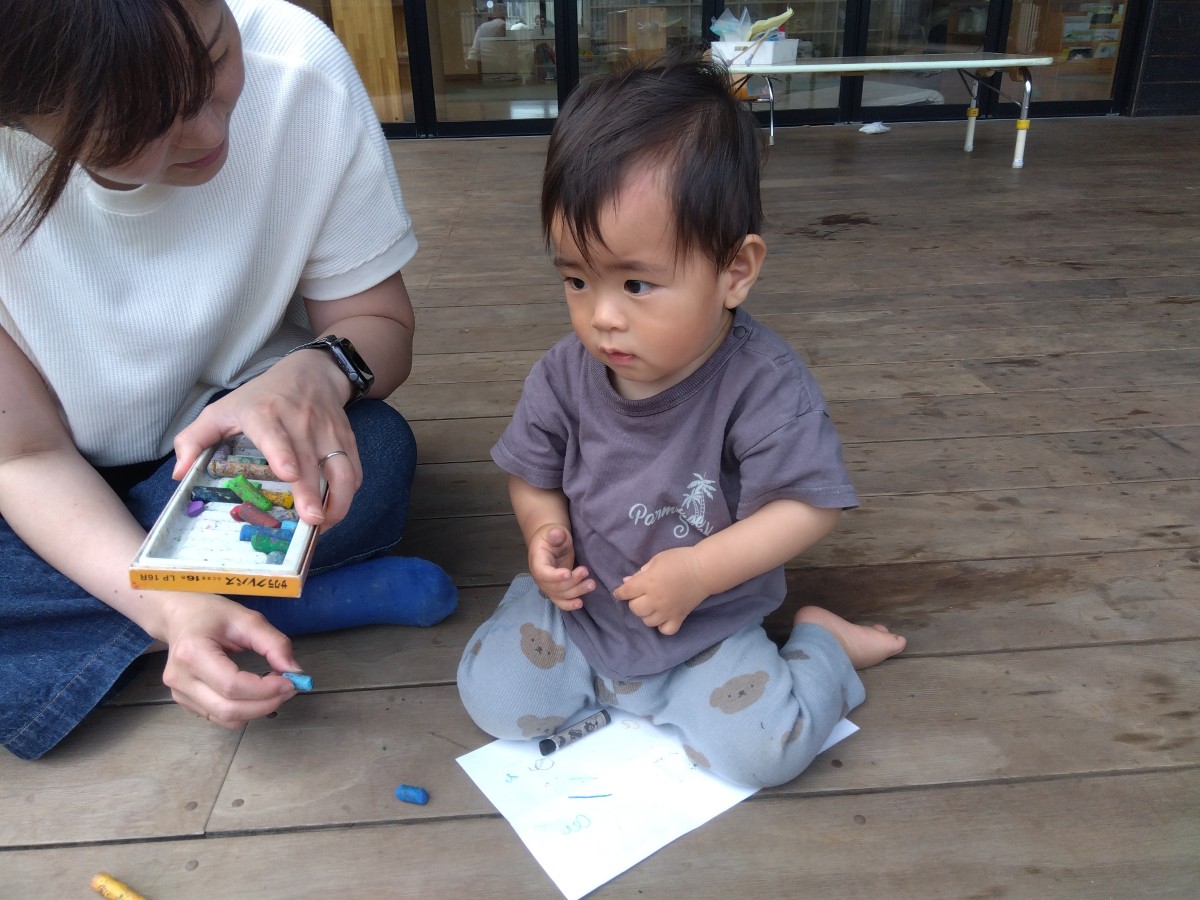 テラスでは親子で並んで座っており、子どもの目の前には画用紙が置かれている。親はクレヨンを子どもに渡そうとしている
