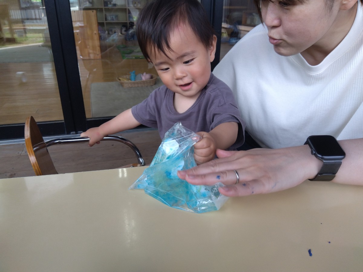 同じ子どもが、洗濯のりや描いた魚が入ったジッパー付きポリ袋を机から持ち上げて握り笑顔を見せている。親は隣でジッパー付きポリ袋を指先で触っている