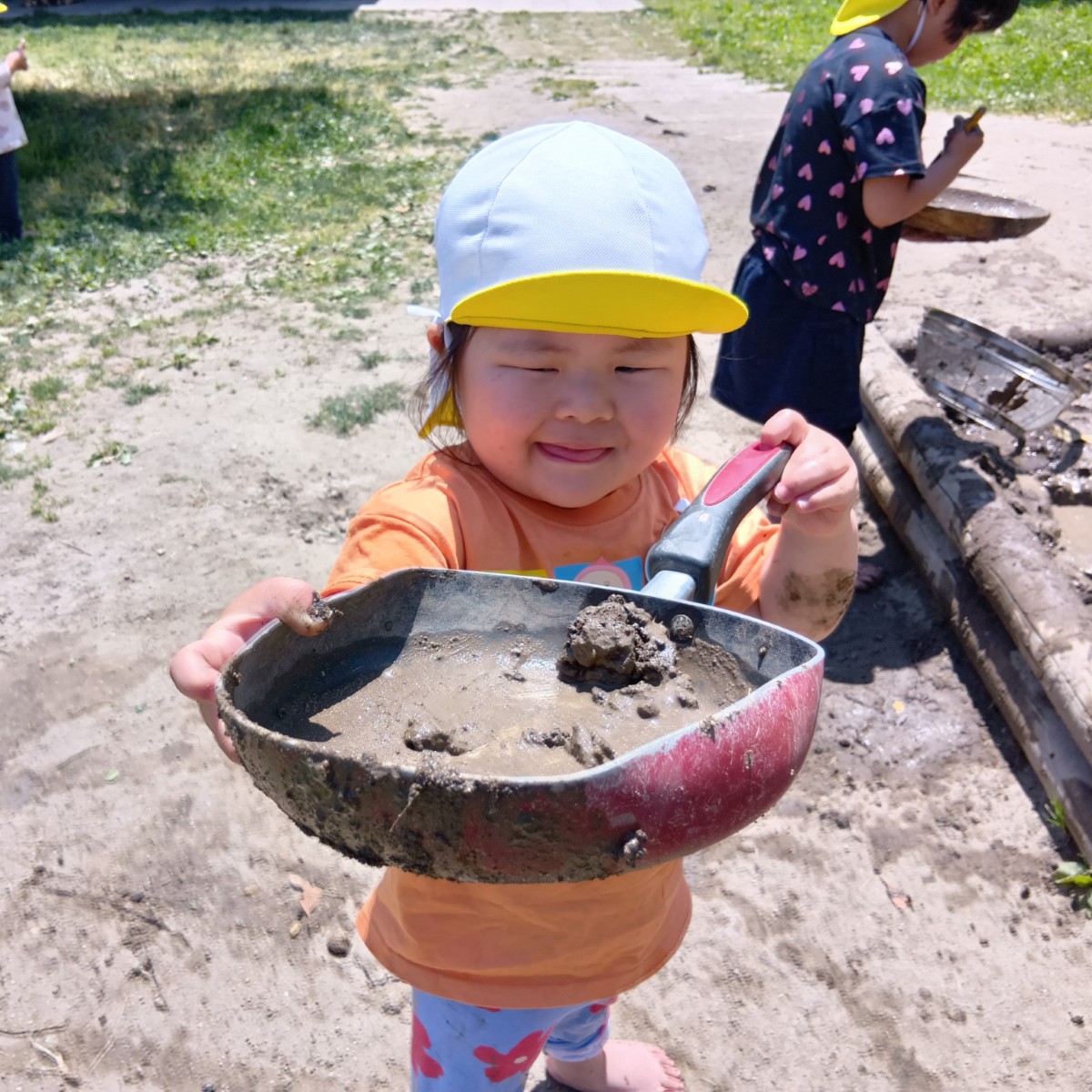 一人の子どもがたくさんの泥が入ったフライパンを両手で持ち上げてこちらに見せている