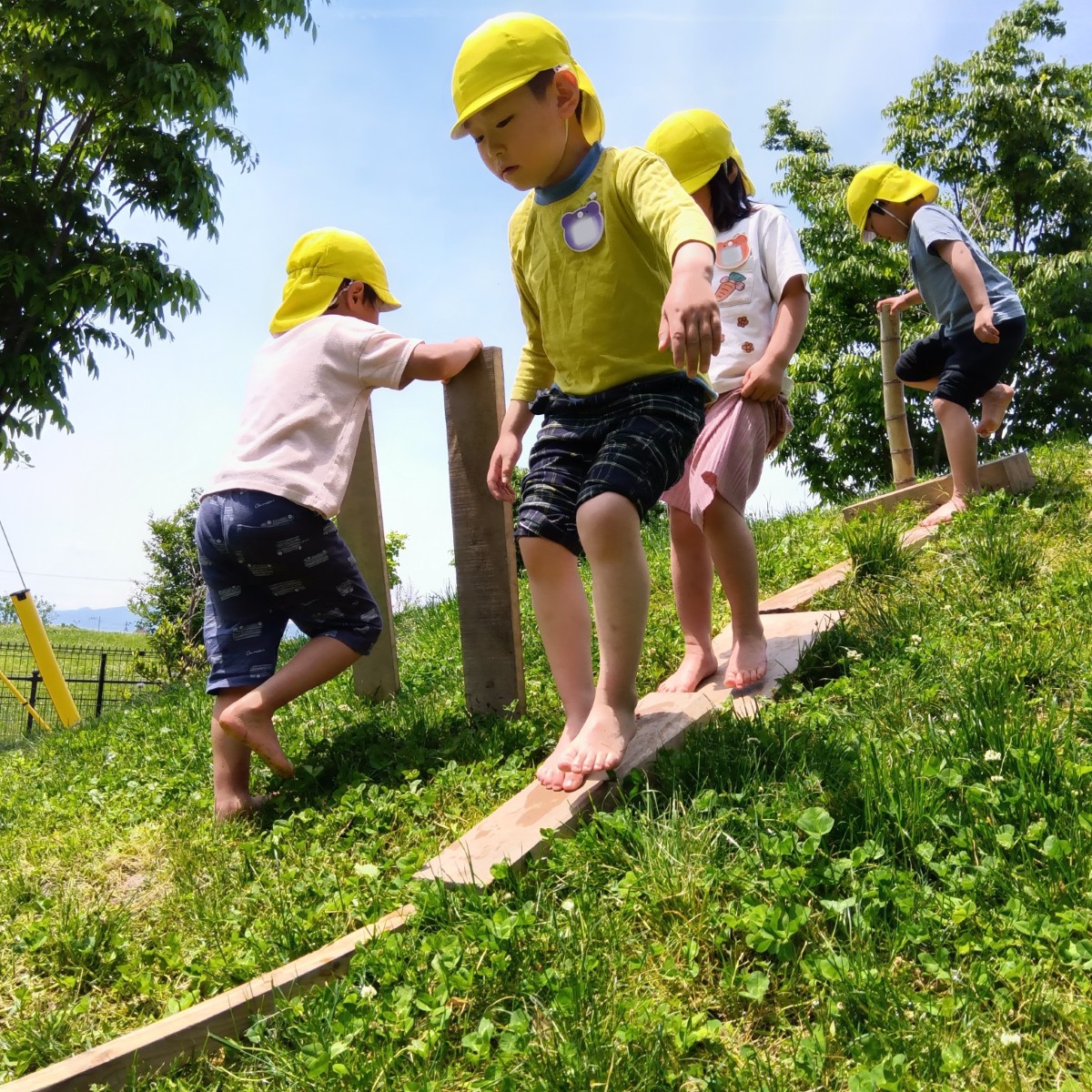斜面に並べられた木材の上を裸足で歩く子、木材を両手に持って傾斜を上る子、竹を持って歩く子などがいる