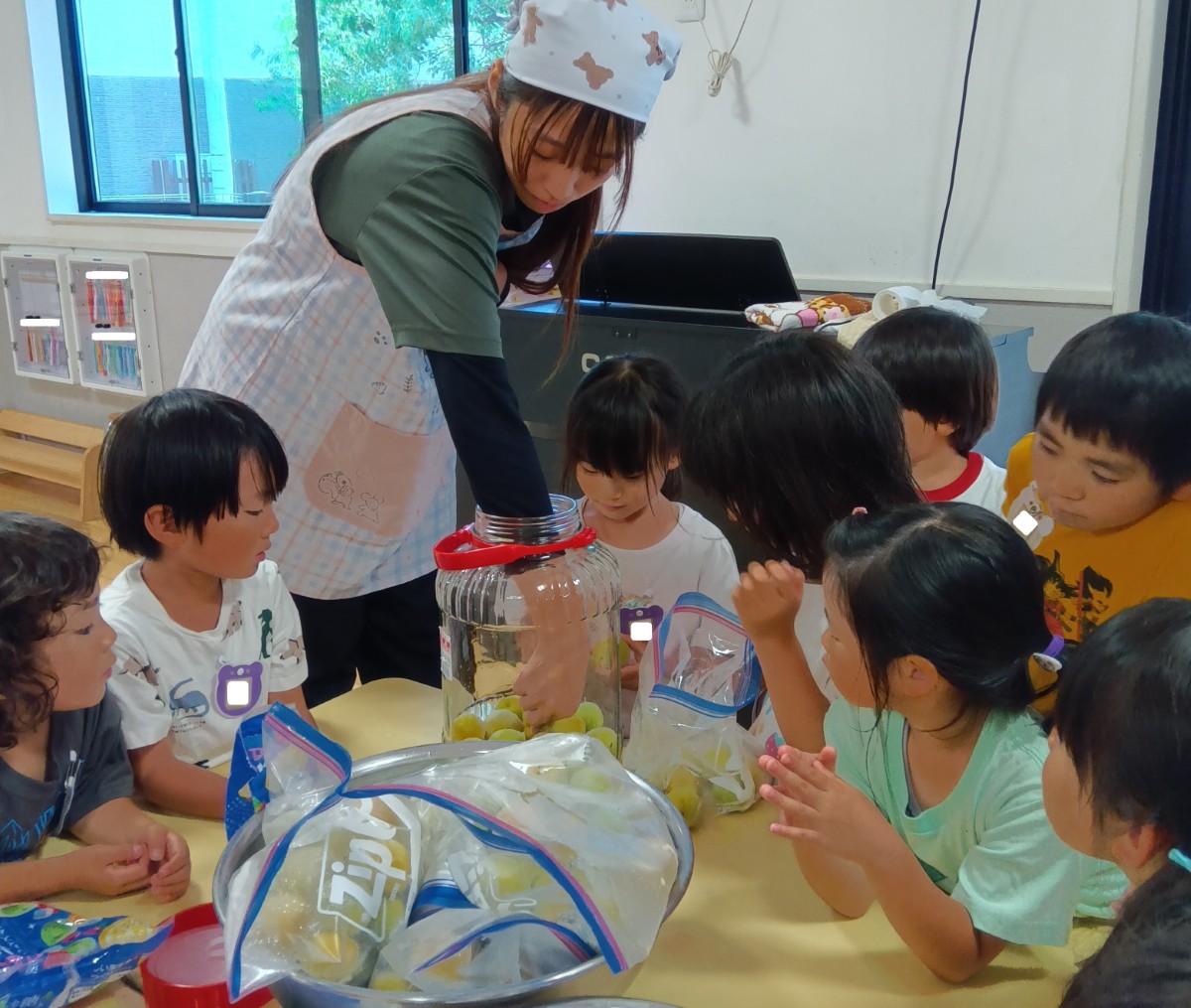 一人の大人が机の上にある瓶に梅を入れており、机の周囲で子どもたちがその様子をみている