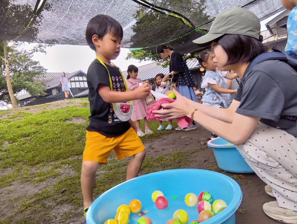 制作で作ったバックを首から掛けたこどもが、水と丸い玩具の入った大きいなたらいから玩具を一つすくって大人に渡している