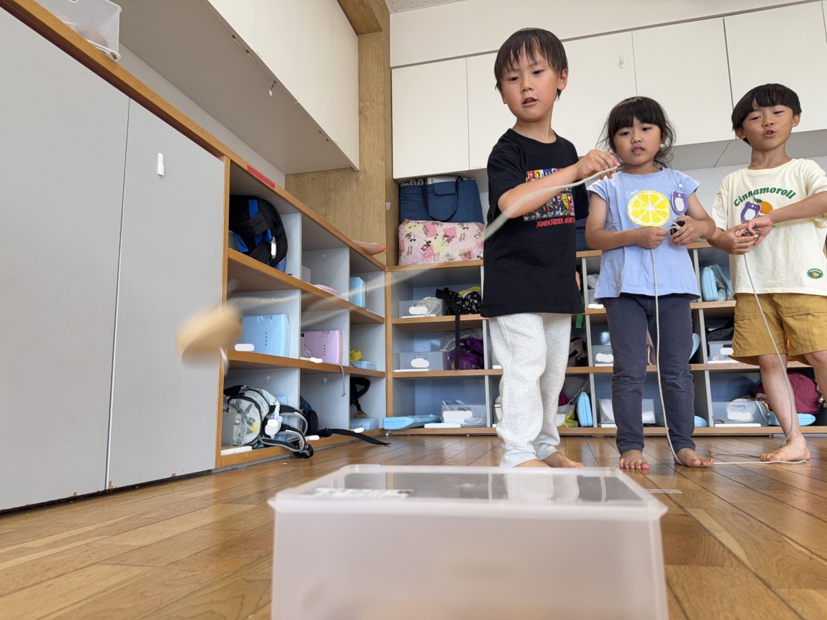 一人の子どもはコマ回し台に向かってコマを投げており、並んでいる子どもたちはコマに紐を巻き付けて順番を待っている