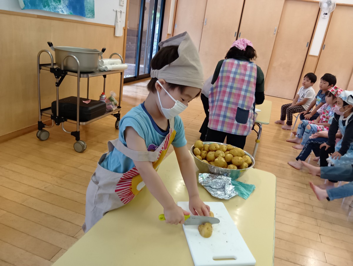 エプロンと三角巾を身に付けた一人の子どもが包丁でじゃがいもを切っており、他の子どもは椅子に座って順番を待っている