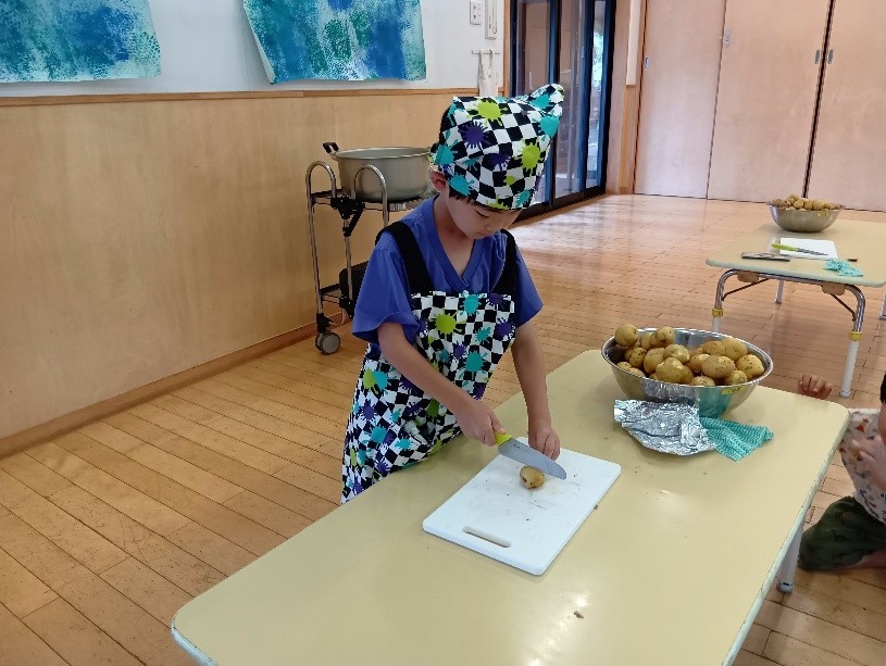 エプロンと三角巾を身に付けた他の子どもも包丁でじゃがいもを切っている