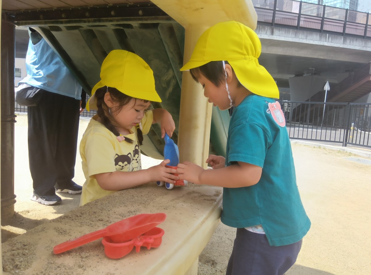 公園では、一人の子どもが砂遊び道具のスコップでカップに砂を入れ、向かいにいる友達とカップの中を見て話している
