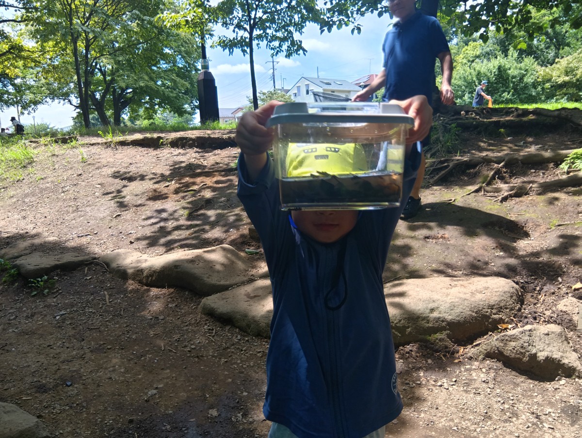 一人の子どもが、水と生き物が入った飼育ケースを顔の前に持ち上げてこちらに見せている