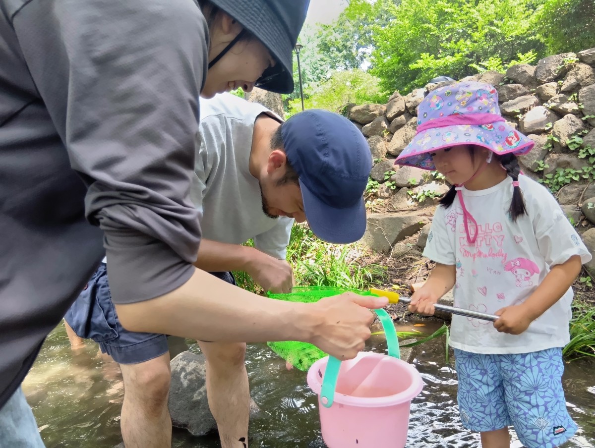 一人の子どもが虫取り網を持ち、二人の大人が網の中の生き物を笑顔で覗き込んでいる