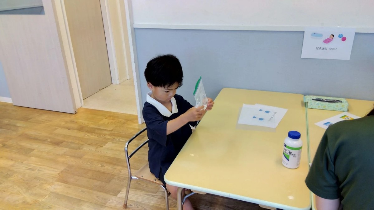 一人の子どもが椅子に座り、両手に持った粉が入った袋を見つめながら揉んでいる