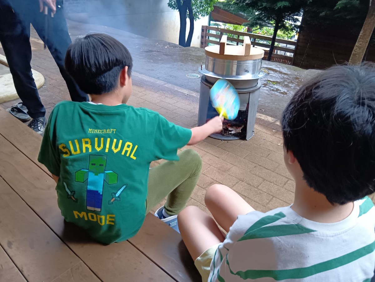 一人の子どもが火起こしした後の釜戸をうちわであおいでいる