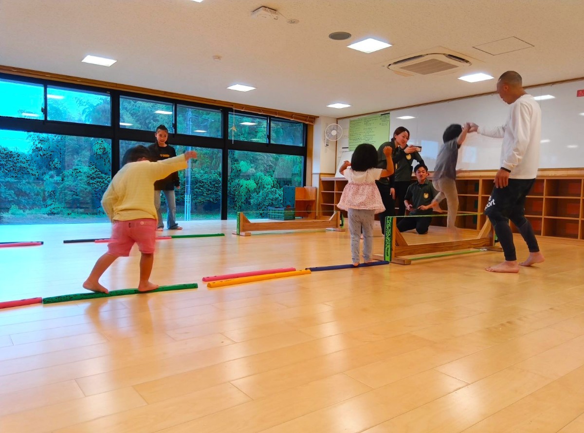 室内では、床に低いウレタン製の平均台が並んでおり、その先にスラックラインが置かれている。子どもたちは低い平均台の上を歩き、大人に手をつないでもらいながらスラックラインを渡っている