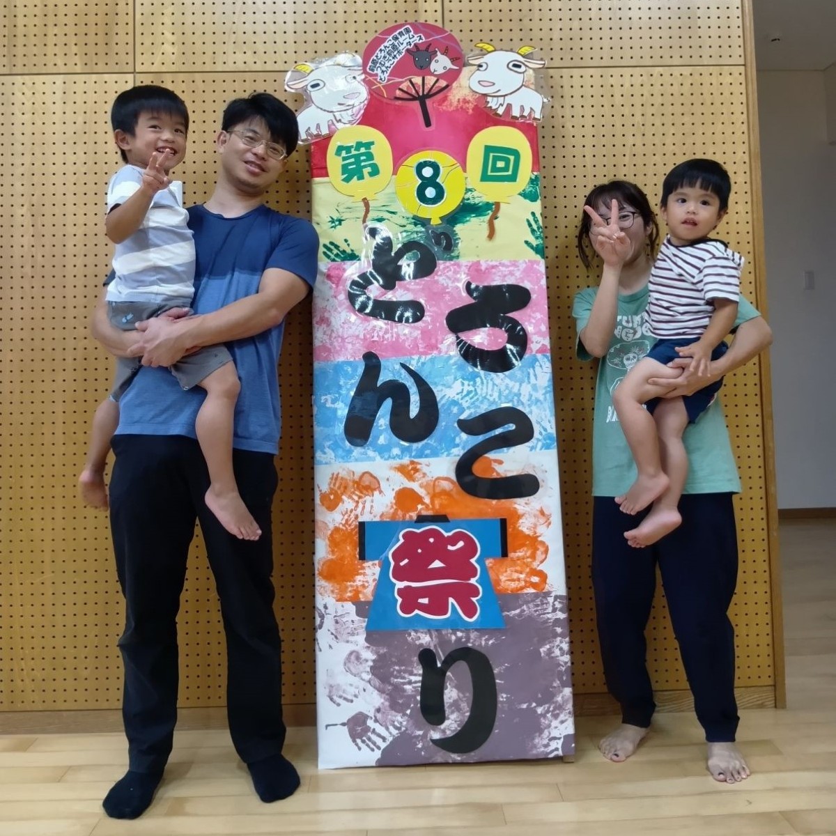 どろんこ祭りの看板の両隣には、子どもを抱えた大人たちが笑顔で立っている