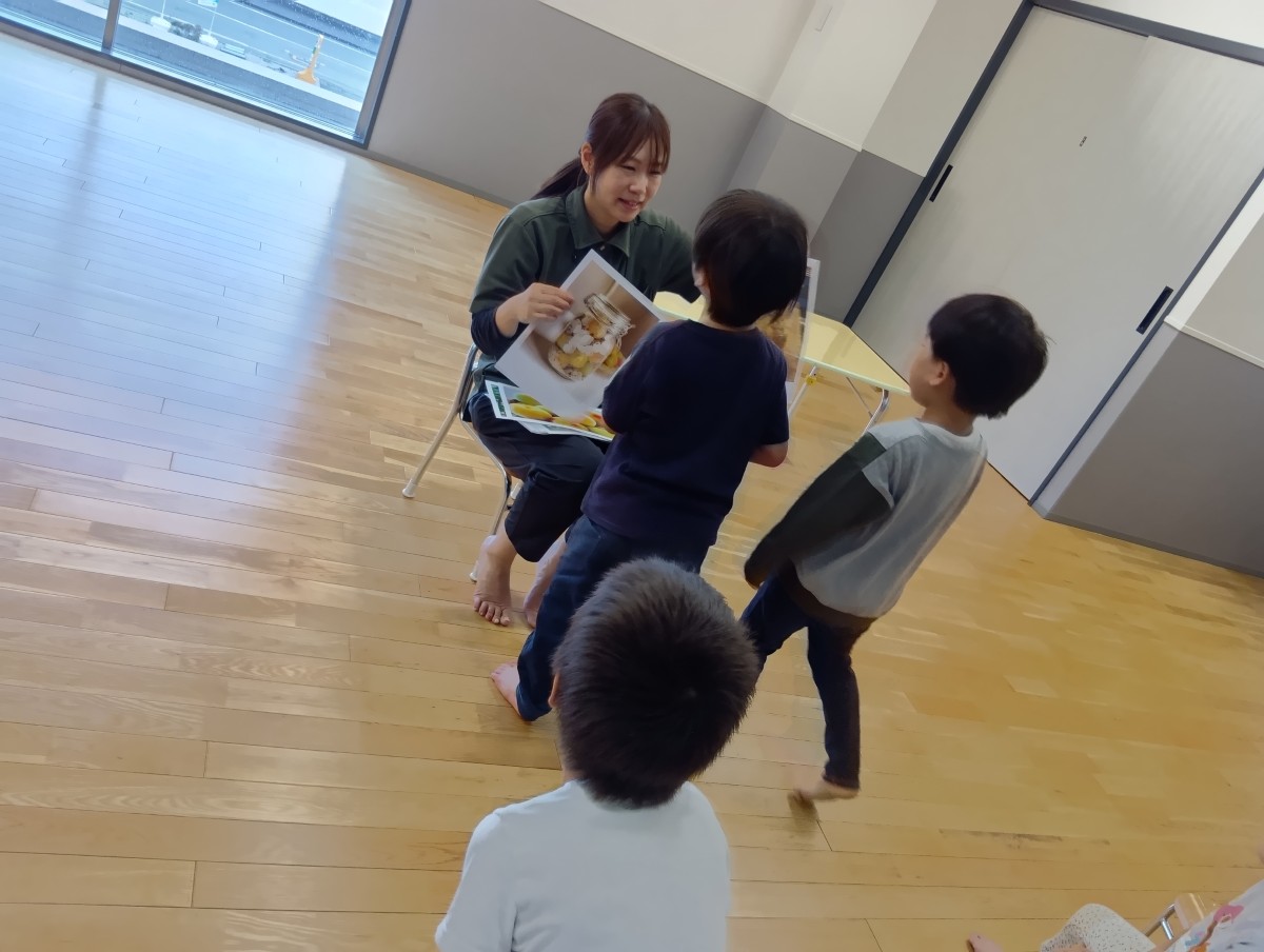 一人の大人が梅が印刷された紙を持って子どもたちの前に座り、二人の子どもが大人の近づいて話しかけている