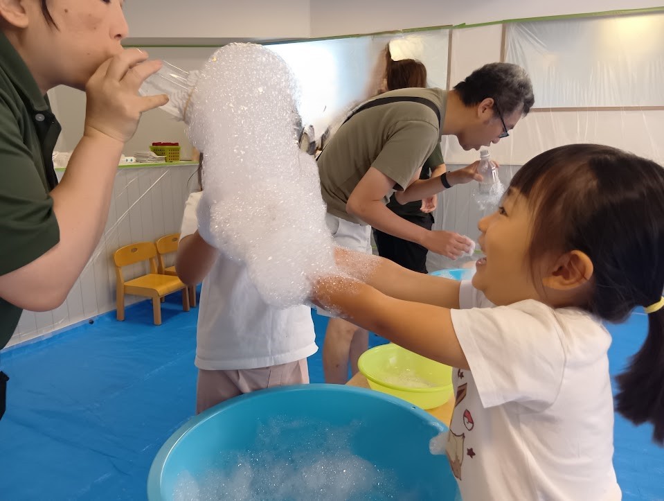 大人が布を巻いたペットボトルに息を吹き込むと、たくさんの泡が出てきて、子どもは喜んで泡に手を伸ばしている