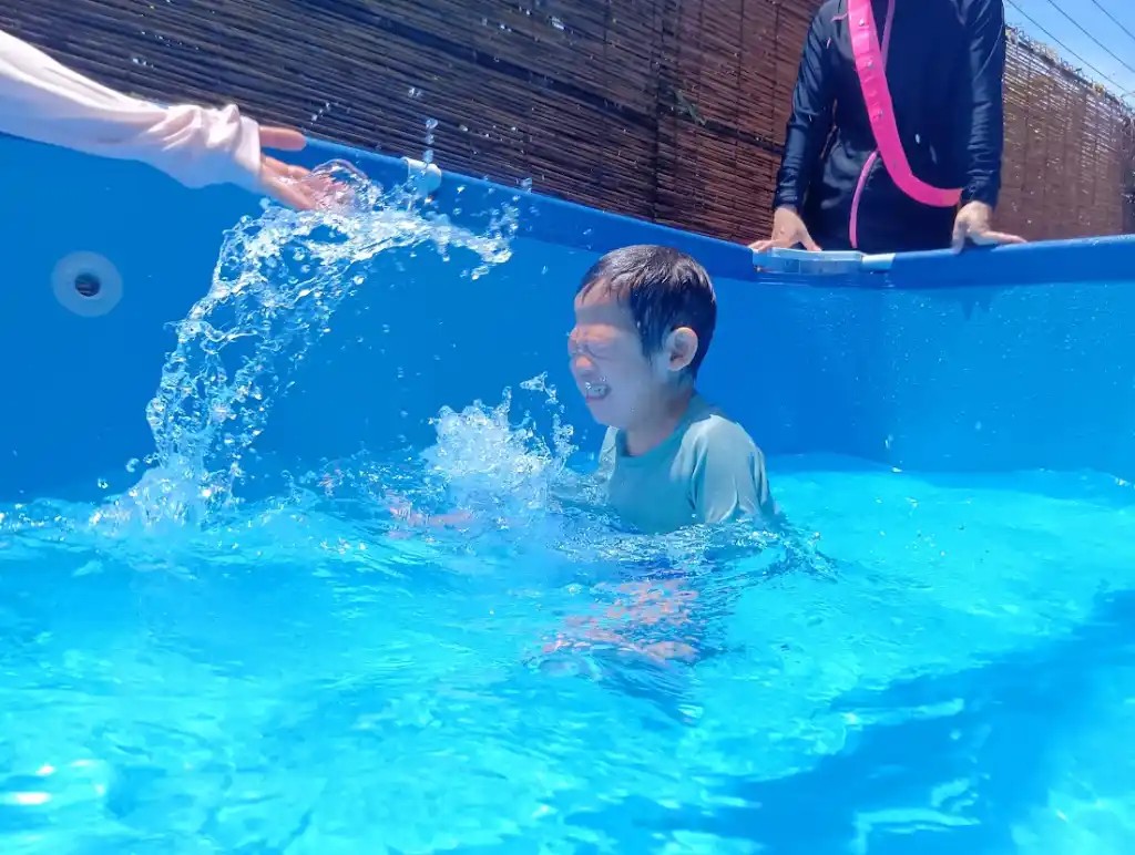 プールの中で、大人が一人の子どもの顔に水をかけると、子どもは目をつぶって喜んでいる。近くでは、もう一人の大人がその様子を見守っている