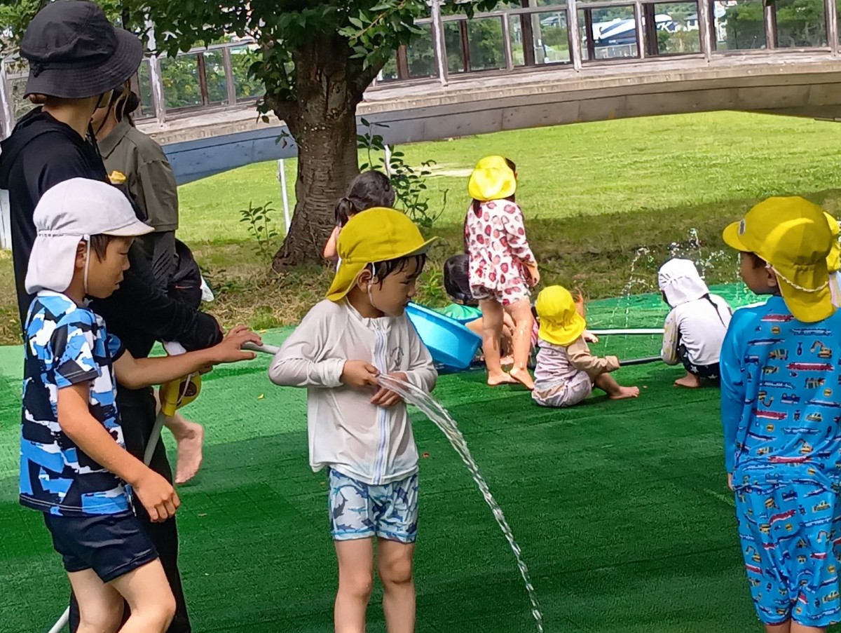 園庭の水遊びでは、地面から出ている水しぶきに集まる子どもたちやホースを自ら持って構える子、それに気が付いて振り返る子などがいる