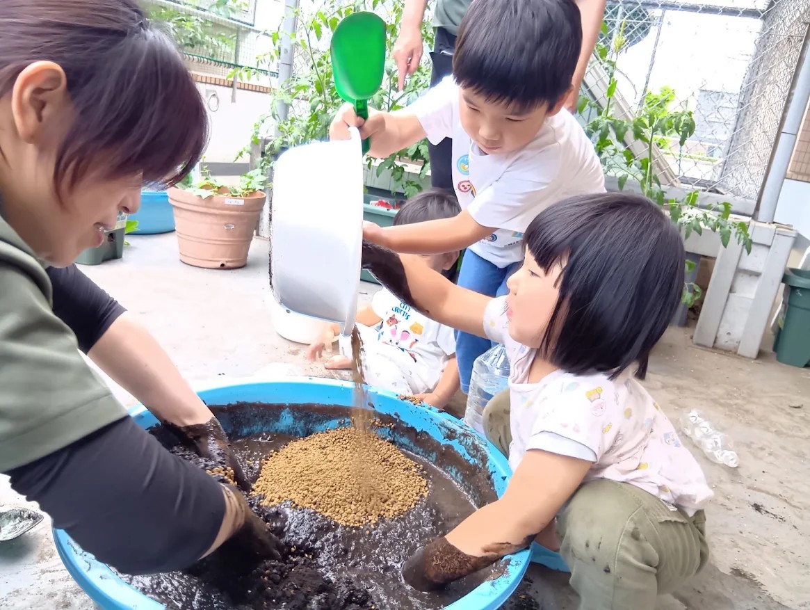 一人の大人と一人の子どもが大きなたらいの中の泥を手で混ぜており、もう一人の子どもはスコップと小さなたらいを持って乾いた土を大きなたらいに入れている