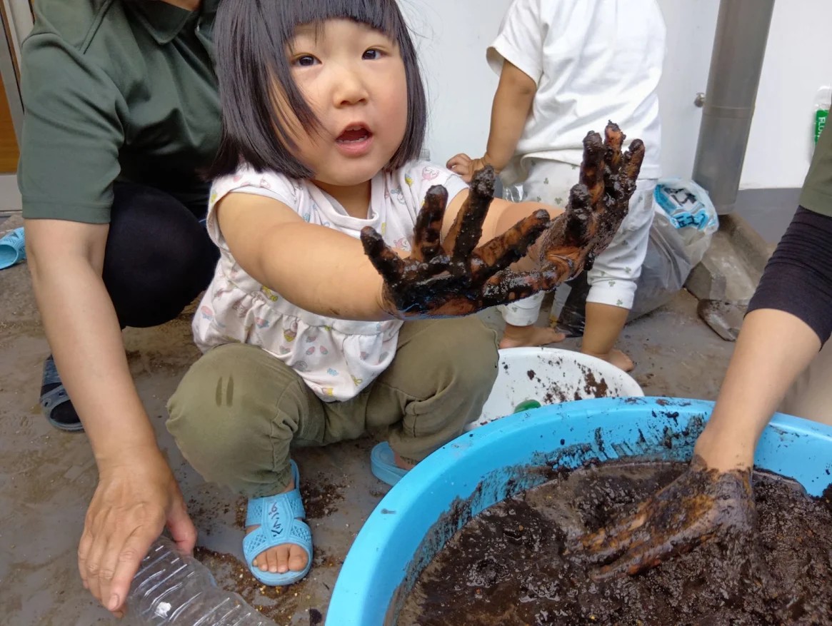 大きなたらいの土を混ぜていた子どもが、泥のついた両手をこちらに見せている