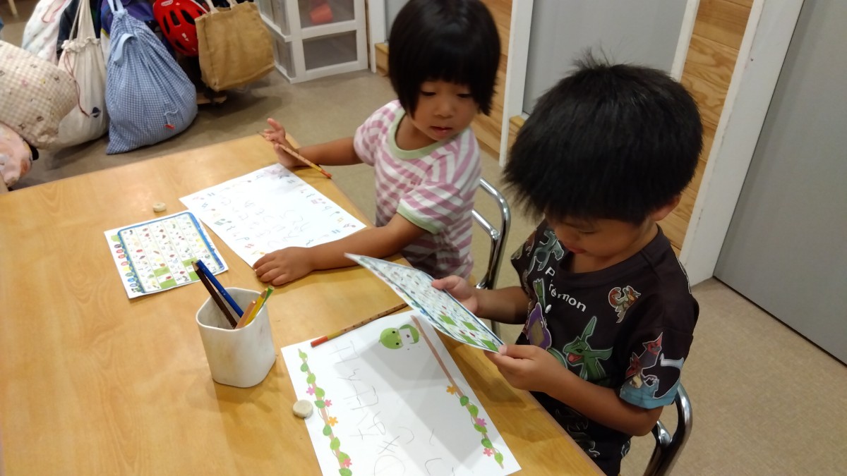 二人の子どもが机に向かい、文字表を見ながら紙に文字を書いている