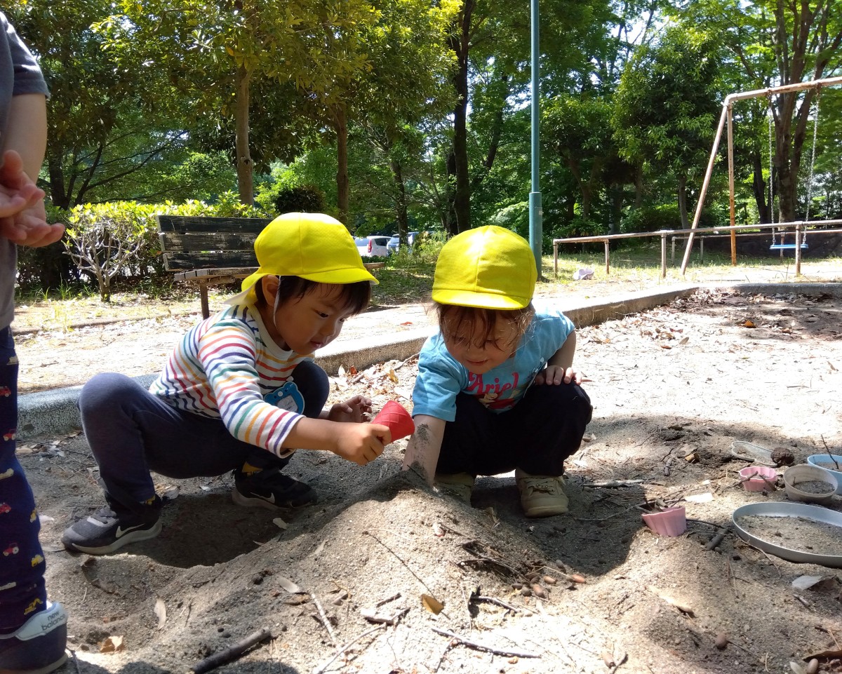公園の砂場では、一人の子どもが砂の中に手を入れており、もう一人の子どもは砂に手を入れている子どもの手にカップで砂をかけようとしている