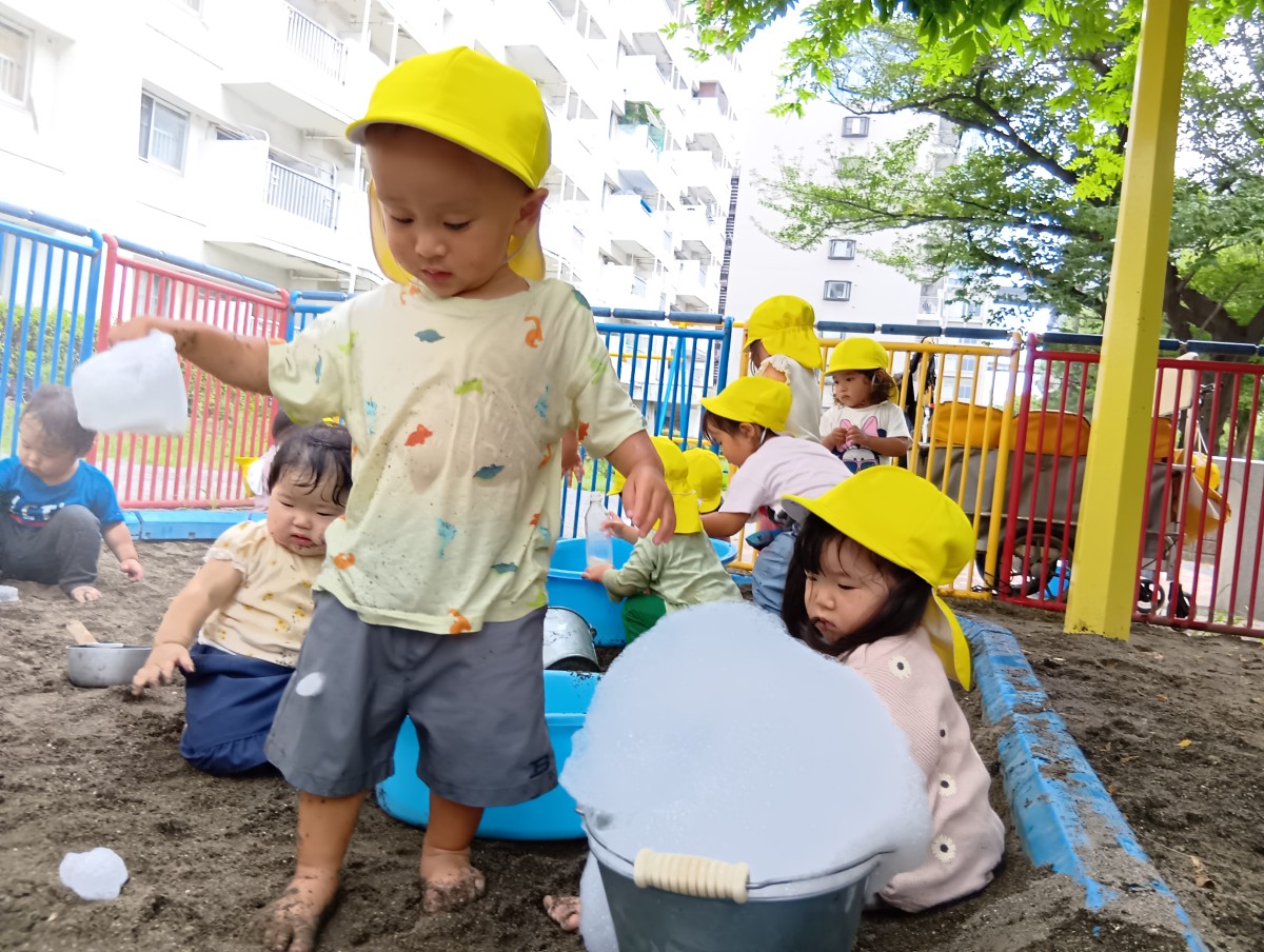 公園では、泡の入ったバケツが置いてあり、たらいに手を伸ばしている子や全身水に濡れている子、ペットボトルに水を入れる子、その様子を見ている子などがいる