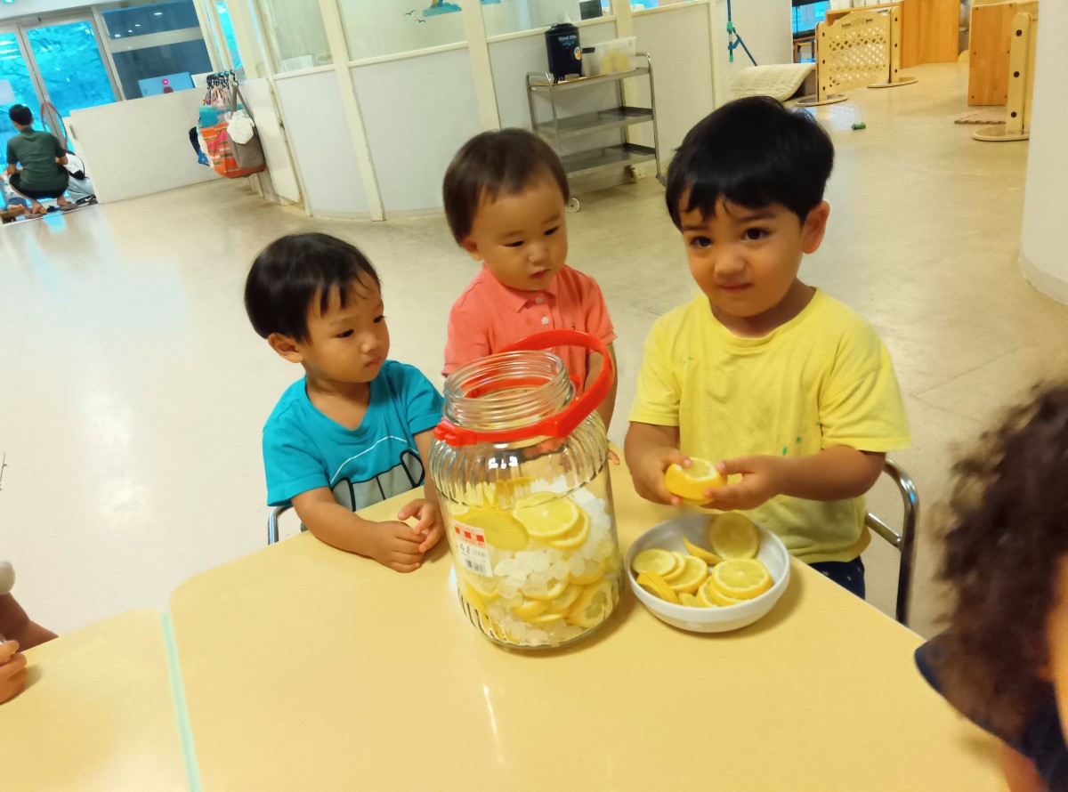 机には輪切りのレモンと氷砂糖が入った瓶が置かれており、一人の子どもがこれから瓶の中に入れる輪切りのレモンを手に持ち、隣には年下児がいる