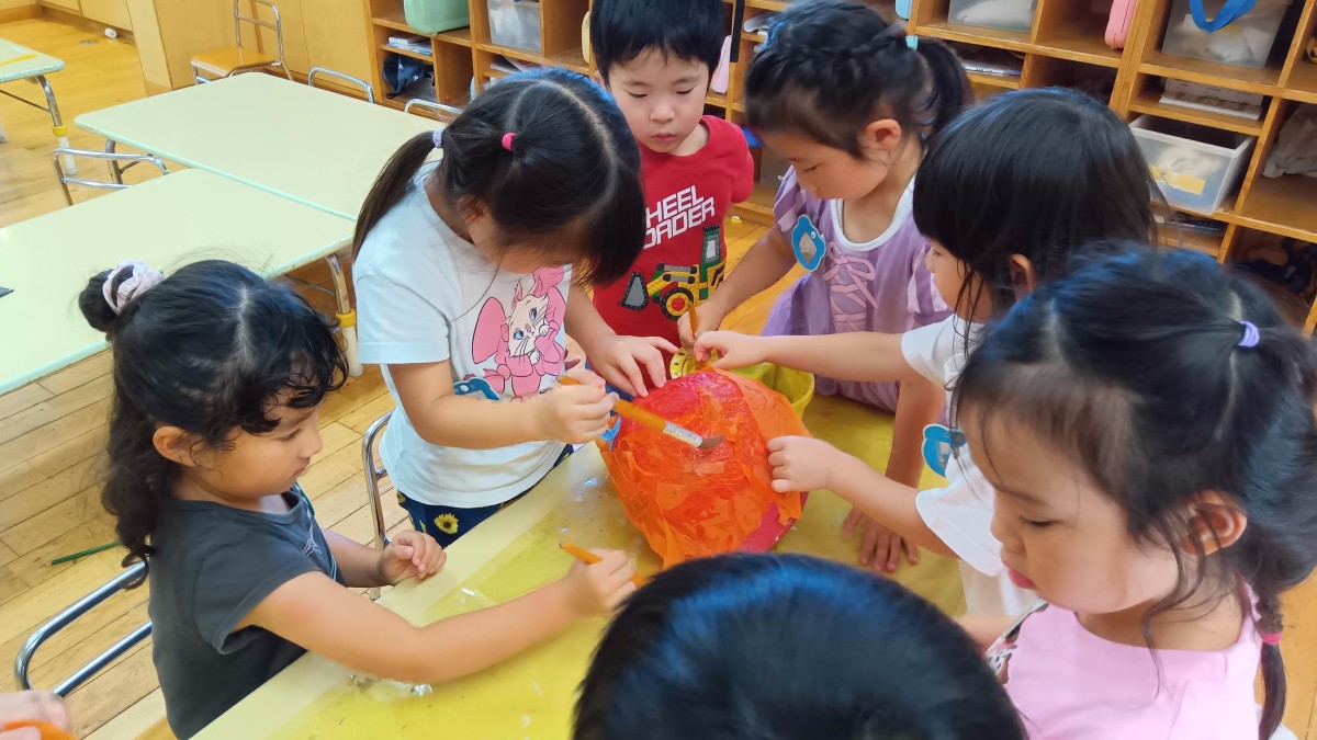 たくさんの子どもたちが机を囲み、提灯を手で支えたり筆で提灯に色を付けたりしている