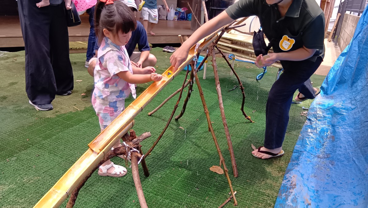 斜めに設置された竹の上をカップゼリーが流れており、それを一人の子どもがスプーンを使ってすくおうとしている