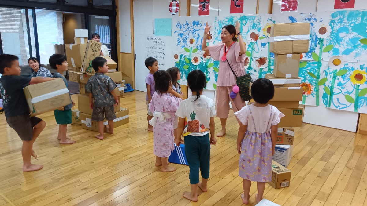 室内では、高く積みあがった段ボールが２つあり、中央に立っている一人の大人が子どもたちに話しかけている