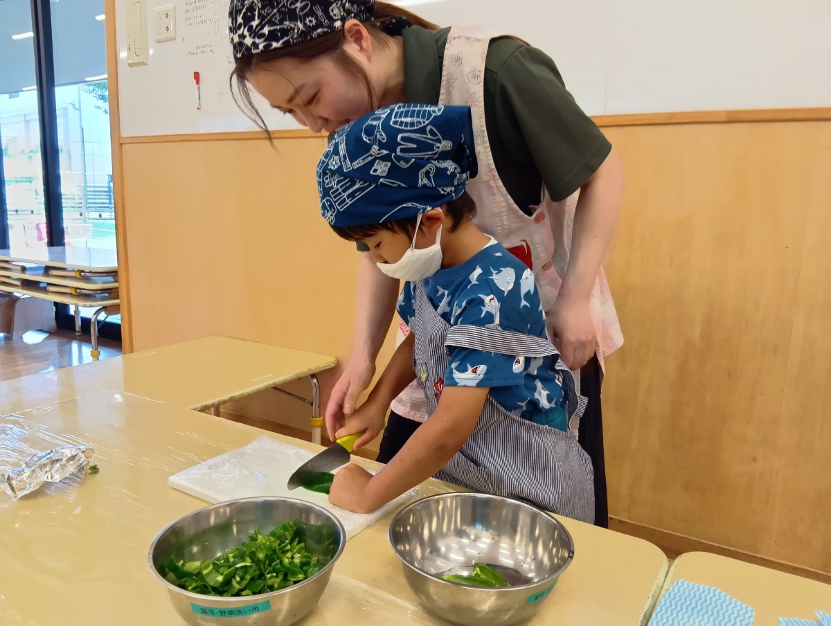 一人の子どもがピーマンを包丁で切っており、子どもの後ろで大人が見守っている