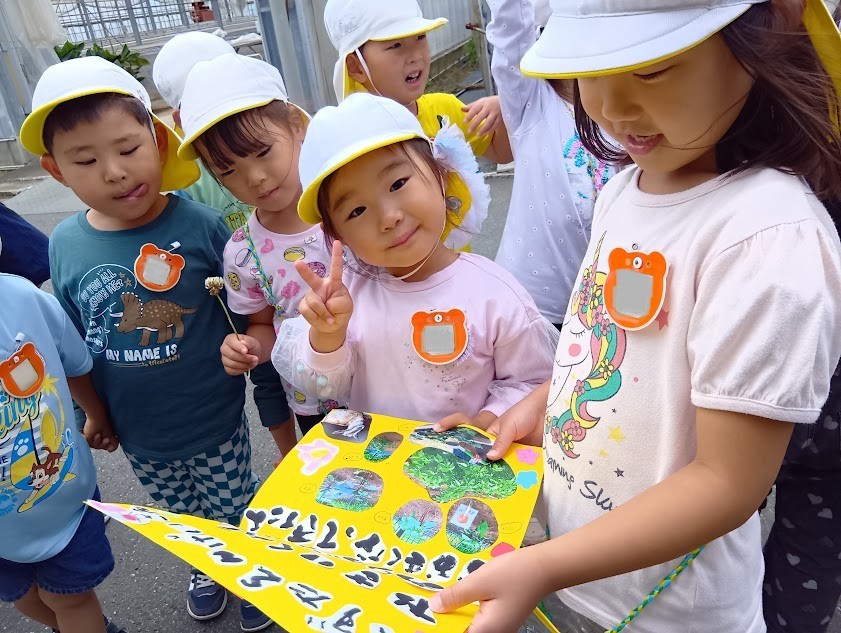 二人の子どもが野菜の成長の写真が貼られた画用紙を持ち、他の子どもたちはその周りを取り囲んで画用紙の中をのぞいている