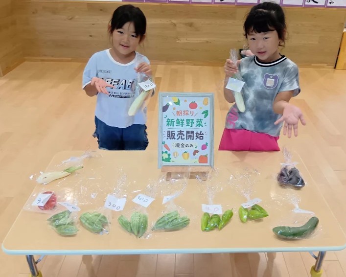 机の上には袋詰めされ値段が貼られた野菜が並んでおり、二人の子どもが販売野菜を手に持っている