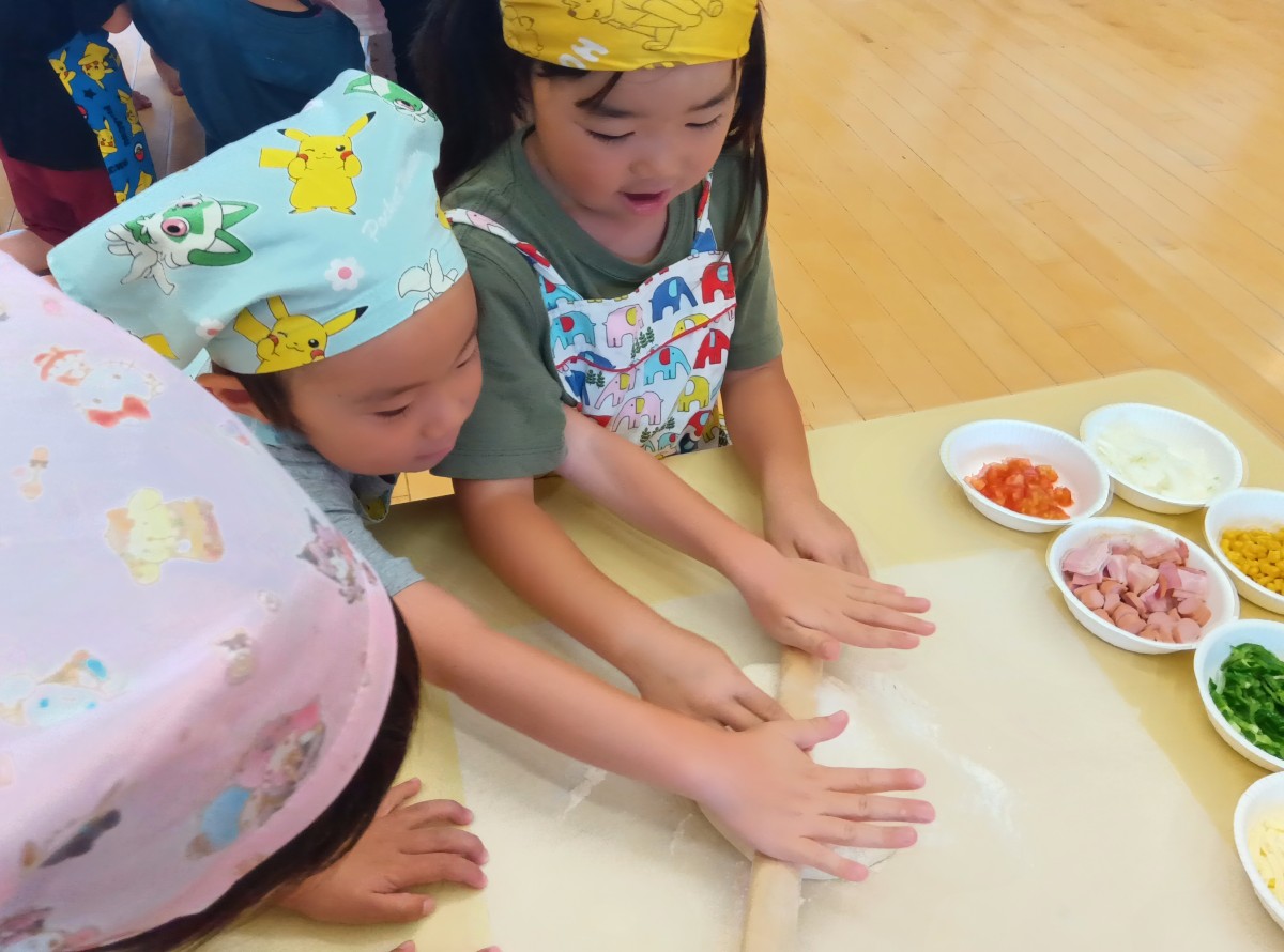 エプロン三角巾を着けた2名の子どもが一緒に麺棒を持ち、机の上のピザ生地を伸ばしている