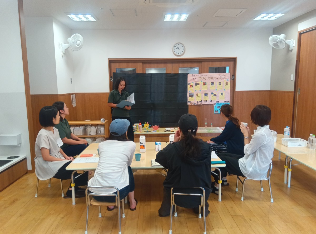 大人たちが机を囲んで前に立つ大人の話を聞いている