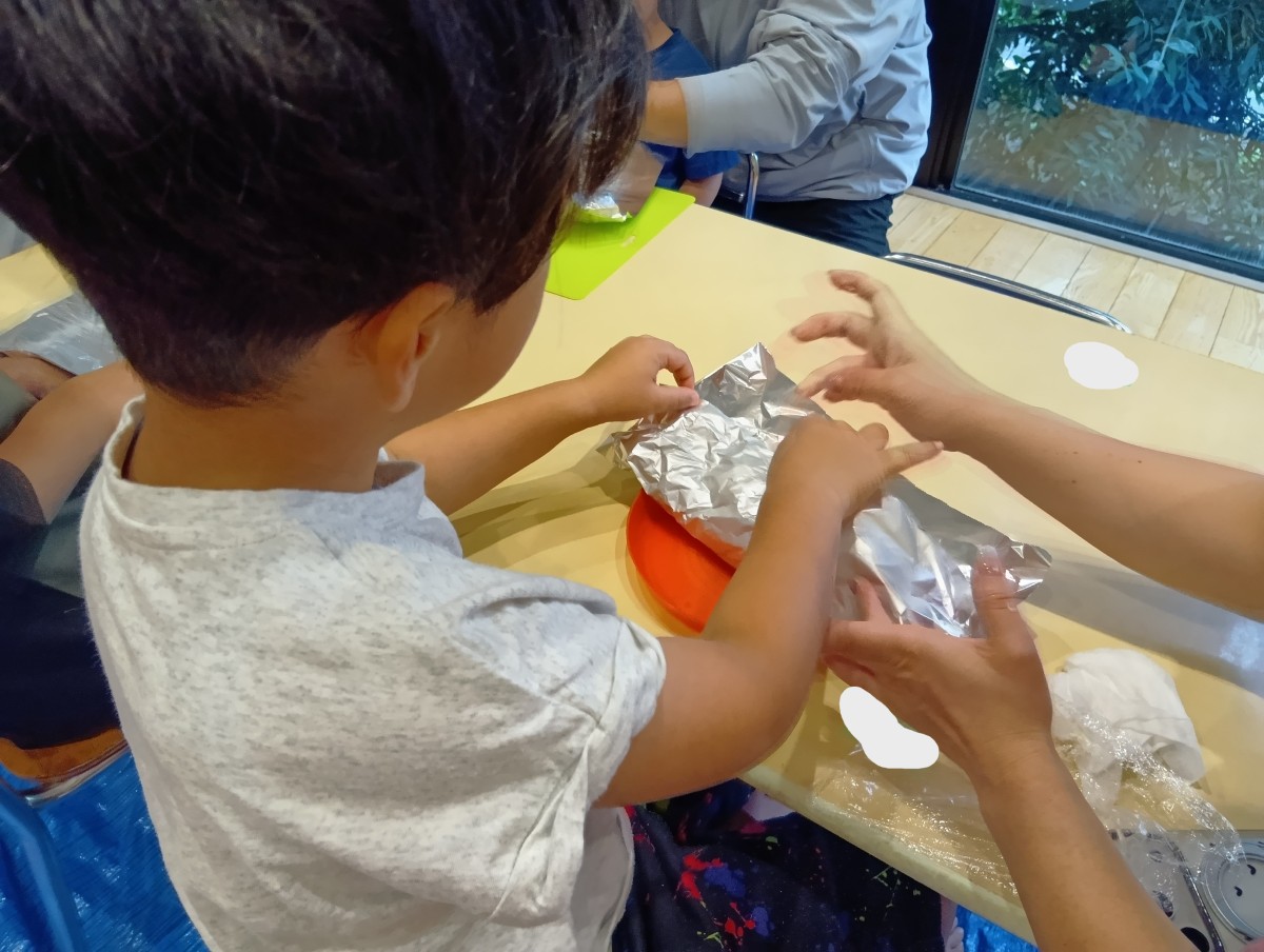 室内の机で、子どもと大人がアルミホイルで生地を包んでいる