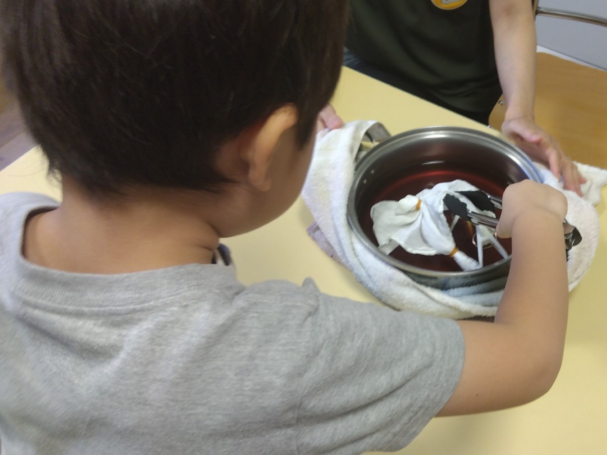 一人の子どもが、鍋に入った染液にゴムで端を留めた布をトングを使って浸している