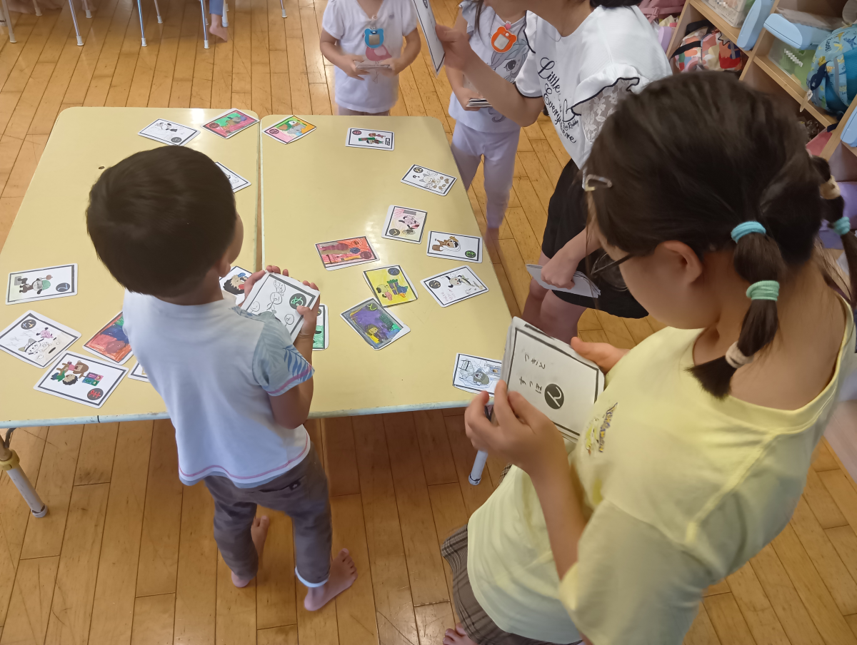 机の上にかるたの取り札が置かれている机の傍には、3名の子どもたちがかるたを手に取って集まり、2名の中学生がかるたを読んでいる