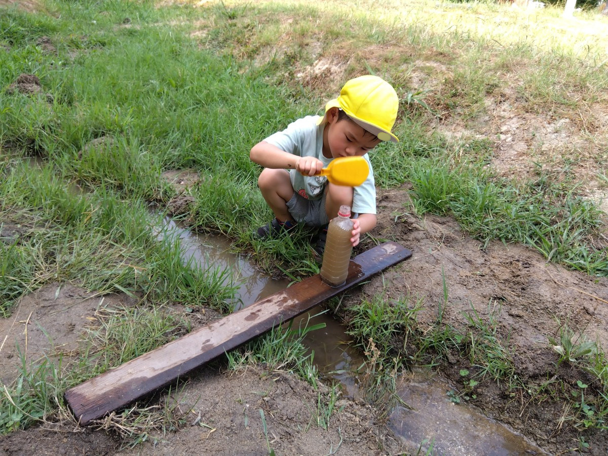 橋が架かっている小川の側で、一人の子どもが橋の上に泥水が入ったペットボトルを置き、シャベルを持ってしゃがんでいる