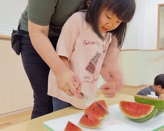一人の子どもがスタッフに手を添えられて、包丁でスイカを切っている