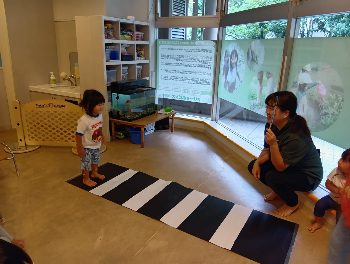 横断歩道が床に敷かれた室内では、大人が信号機を手に持ち子どもは歩道を渡ろうとしている